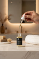 Bottle of skincare serum with a brush, soap, and scrubber on a tiled bathroom counter.