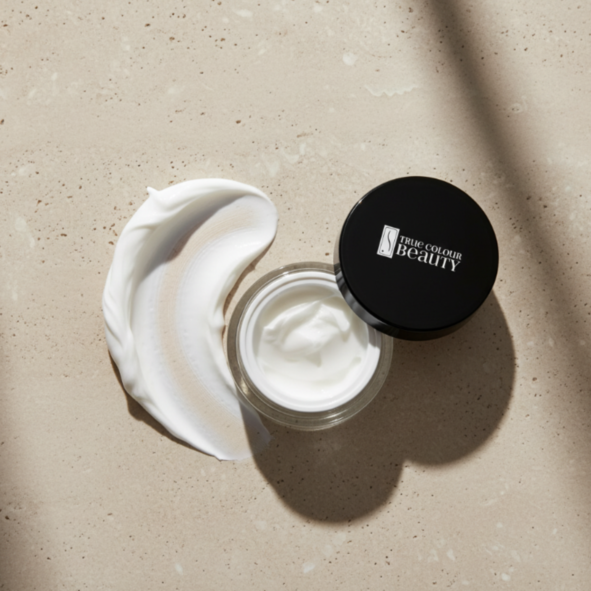 Open jar of cream with a swatch on a beige surface, featuring 'True Colour Beauty' brand.