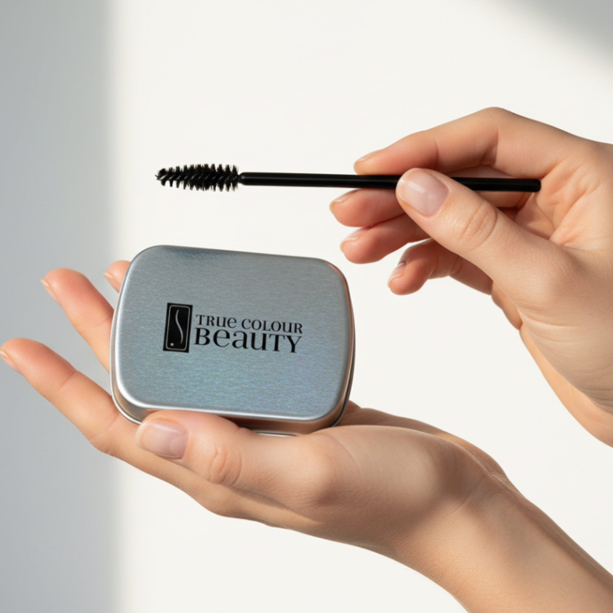 Hand holding a compact mirror with 'True Colour Beauty' logo and a mascara wand.