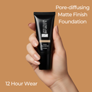 Hand holding a black foundation tube with text about pore-diffusing matte finish foundation on a brown background
