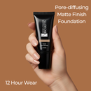 Hand holding a black foundation tube with 'The Colour Beauty' branding on a brown background.