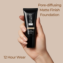Hand holding a black foundation tube with text about pore-diffusing matte finish foundation on a beige background