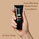 Hand holding a tube of foundation with text about pore-diffusing matte finish and 12-hour wear on a beige background