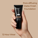 Hand holding a black foundation tube with text about pore-diffusing matte finish foundation on a beige background