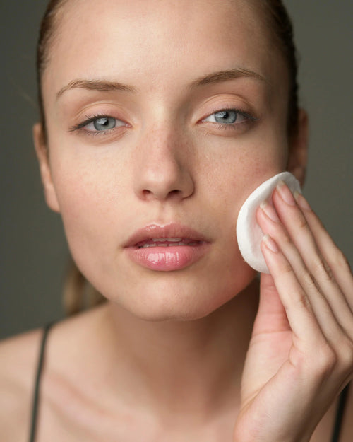 Woman applying a facial product with a cotton pad against a neutral background