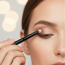 Person applying makeup with a brush to their eye, blurred lights in the background