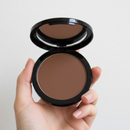 Hand holding a compact of brown powder with a mirror on a light background