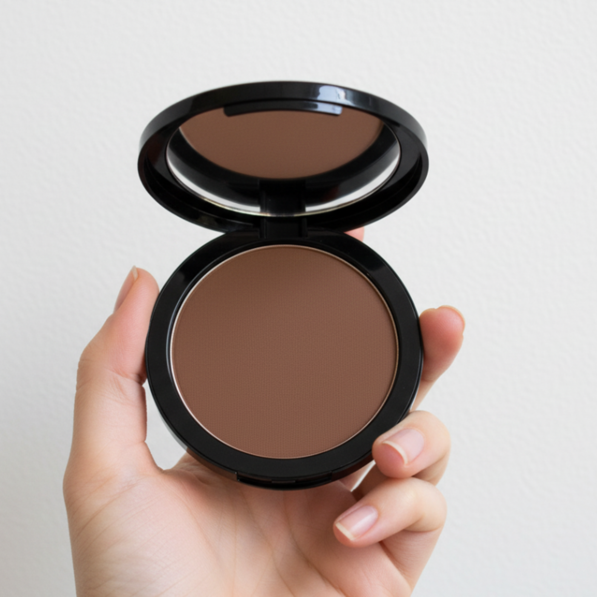 Hand holding a compact of brown powder with a mirror on a light background