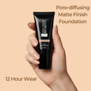 Hand holding a black foundation tube with text about pore-diffusing matte finish and 12-hour wear on a beige background