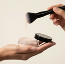 Person holding a makeup brush and jar of powder against a beige background